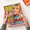 Titelbild der Zeitschrift „Freizeit Revue“ mit großem Porträt einer blondhaarigen Frau auf blaugrünem Hintergrund. Textaufschriften u. a.: „Maite Kelly – Neues Glück mit einem Rosenkavalier“, „Not-OP am Herzen“, „Peinliche Liebesshow“, „Süße Schwärmerei“. Gelbes, rotes und weißes Schriftlayout, kleinere Fotos mehrerer Personen, Zeitschrift wird mit beiden Händen über hellem Tisch und orangem Tablett gehalten.