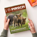 Magazin „Pirsch“ Ausgabe 11/2025 mit braunem Titelbalken und weißer Schrift „PIRSCH – Respekt vor dem Wilden. Seit 1879“. Titelbild zeigt zwei Wildschweine, eines im Vordergrund auf grüner Wiese, das andere verschwommen im Hintergrund. Weitere sichtbare Texte: „Rehböcke“, „Aufbrechsägen“, „Sommersauen – Das müssen Sie bei der Nachtjagd beachten“. Gehalten von zwei Händen über hellem Tisch mit orangefarbenem Objekt oben rechts.