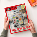 Magazin „FOCUS“, Ausgabe 18 vom 24. April 2024, mit rotem Rand und Titelmotiv eines silbernen Rollkoffers, beklebt mit Stickern u. a. von Flugzeug, Geldschein, Weltkugel, rotem Auto und Symbolfiguren; Überschrift in Weiß und Rot: „ANGST UM DEN URLAUB – Die 30 wichtigsten Fragen zum Krisen-Sommer 2026“, weitere Textzeilen zu Migration und E‑Bike‑Spezial, gehalten von zwei Händen über heller Tischoberfläche.