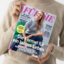 Person in beigefarbenem Strickpullover hält das Magazin „FÜR SIE“ mit einer lächelnden Frau auf dem Cover, die in grauem Pullover und dunkelblauer Hose auf einem grünen Stuhl sitzt. Sichtbare Texte: „Die Formel für ein langes Leben“, „Healthy Aging: Simple Tipps für einen gesunden Körper, ein fittes Gehirn und mehr Lebensfreude“, sowie Schlagzeilen zu Farben, Mustern, Beziehungen, Reisen und Gesundheit.