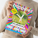 Person in beigem Strickpullover hält die Zeitschrift „happinez – Das Mindstyle Magazin“ mit bunt gestreiftem, kreisförmigem Coverdesign. Zentrale Illustration: eine Person im Handstand auf einer grünen Wiese mit Blumen und Bäumen. Sichtbare Texte: „Sei glücklich – Alles strahlt heller, wenn Du lachst“, „EXTRA Zauberhafte Lesezeichen“, „Selbst-Erkenntnis“, „Liebes-Magie“, „Aktive Hoffnung“, „Weiblichkeit – Die Superkraft Deines Zyklus“.