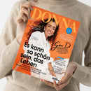 Person in beigem Strickpullover hält das Magazin „DONNA“ mit orangefarbenem Titelblatt hoch; darauf eine lächelnde Frau mit lockigem, dunklem Haar in weißer Bluse. Hauptaufschrift: „Es kann so schön sein, das Leben“. Weitere Texte: „Gen D. Generation DONNA – was sich für Frauen in den Wechseljahren ändern muss“, „Amore! Italienische Rezepte, ganz easy“ und Zitat „Ich will nicht jünger aussehen“.