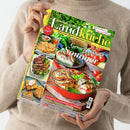 Titelblatt der Zeitschrift „meine gute Landküche“ mit dem Haupttext „Genießen im Sommer“. Abgebildet sind ein roter runder Auflauftopf mit Gemüse-Quiche in Sahnecreme, frische Tomaten und Kräuter im Hintergrund sowie kleinere Fotos mit Focaccia, Steaks, Spießen und Kuchen. Eine Person in beigem Pullover hält das Magazin vor neutralem Hintergrund in beiden Händen.