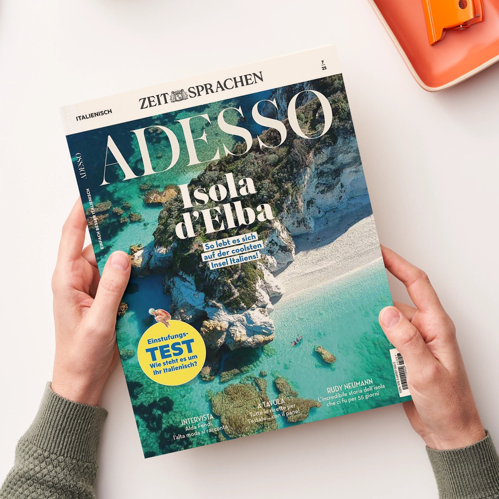 Titelblatt des Magazins „Adesso“ der Reihe „Zeit Sprachen“, Ausgabe 7/25, mit der Überschrift „Isola d’Elba – So lebt es sich auf der coolsten Insel Italiens!“. Das Cover zeigt eine Luftaufnahme der Küste von Elba mit türkisblauem Wasser, hellen Felsen und einem Strand. Unten links gelber Kreis mit der Aufschrift „Einstufungs-Test: Wie steht es um Ihr Italienisch?“. Gehalten von zwei Händen über hellem Hintergrund.