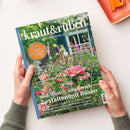 Titelblatt der Zeitschrift „kraut&rüben“ mit grün-weißem Schriftzug und Unterzeile „Schöner. Wilder. Biogarten.“; im Hintergrund blühender Garten mit rosa und weißen Rosen, grünem Laub und unscharfem Gebäude mit Fensterläden. Sichtbare Texte: „Großes Sommer-Gewinnspiel“, „Wild, sinnlich, sommerlich – Gestalten mit Rosen“, „Sauerkirschen“, „Schneckenfeste Blütenstauden“. Zwei Hände halten die Ausgabe auf hellem Tisch.