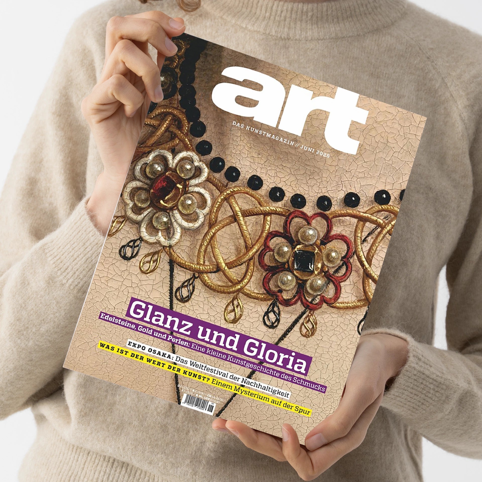 Person in beigem Strickpullover hält mit beiden Händen die Zeitschrift „art – Das Kunstmagazin / Juni 2023“. Titelbild zeigt eine goldene Schmuckverzierung mit roten, schwarzen und perlenfarbenen Elementen auf strukturierter Oberfläche. Sichtbare Textzeilen: „Glanz und Gloria“, „Edelsteine, Gold und Perlen: Eine kleine Kunstgeschichte des Schmucks“, „EXPO OSAKA“ und „WAS IST DER WERT DER KUNST?“.