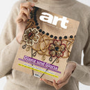 Person in beigem Strickpullover hält mit beiden Händen die Zeitschrift „art – Das Kunstmagazin / Juni 2023“. Titelbild zeigt eine goldene Schmuckverzierung mit roten, schwarzen und perlenfarbenen Elementen auf strukturierter Oberfläche. Sichtbare Textzeilen: „Glanz und Gloria“, „Edelsteine, Gold und Perlen: Eine kleine Kunstgeschichte des Schmucks“, „EXPO OSAKA“ und „WAS IST DER WERT DER KUNST?“.