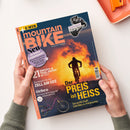 Titelbild des Magazins „mountainBIKE“ mit orange-gelbem Sonnenuntergang im Hintergrund und einer Person auf einem Mountainbike in dunkler Silhouette im Vordergrund. Aufschriften u. a.: „Der Preis ist heiß“, „21 Enduro- & Trail-Reifen im Test“, „Zell am See“, „Orbea“, „Shimano XT Di2 im Stresstest“ und „Neu: 16 Seiten Community“. Zwei Hände halten das Heft über einer hellen Tischoberfläche.