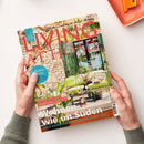 Magazintitel „LIVING AT HOME“ in großen roten Buchstaben über einem Foto einer mediterranen Terrasse mit Steinwand, grünem Sofa, Beistelltisch und Dekoobjekten. Sichtbare Texte: „Wohnen wie im Süden“, „Urlaubsreiche“, „Sommerliche Ideen für den Tisch, Geschirr, Stoffe, Accessoires & DIY“, „Citytrip München“. Gehalten von zwei Händen, Hintergrund in hellem Beige mit orangefarbenem Tablett oben rechts.