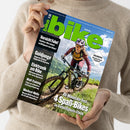Person in beigefarbenem Pullover hält das Magazin „bike“ (Ausgabe 06/2023) in beiden Händen, Titel in leuchtendem Grün auf blauem Himmel-Hintergrund. Auf dem Cover eine weibliche Radfahrerin mit Helm und Sonnenbrille auf einem Mountainbike im Gelände, grüne Berge im Hintergrund. Sichtbare Schlagzeilen: „Vorsicht Fake!“, „Goldjunge“, „Elektronik am Bike“, „Watt-Training“, „Wechsel-Welle“, „4 Spaß-Bikes“, „Dolomiten“.