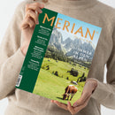 Eine hellhäutige Person in beigem Strickpullover hält das Magazin „MERIAN – The Art of Travel“ in den Händen; Titelthema „Sommer in den Alpen“ mit Unterzeile über Wandererlebnisse in Salzburg, Tirol und der Schweiz. Auf dem Cover eine Fotografie mit grünen Almen, einer braun-weißen Kuh und Bergen im Hintergrund; linke Spalte mit weiteren Themen: „Mexico City“, „Korfu“, „München isst so gut!“ und „Teneriffa“.