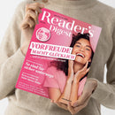 Beige gekleidete Person hält das Magazin „Reader’s Digest Deutschland“ (Ausgabe Juni 2023) mit pinkem Titelblatt, auf dem eine lächelnde weiblich gelesene Person in rosa Oberteil zu sehen ist; sichtbare Überschriften u. a. „Vorfreude macht glücklich“, „So sind Sie sicher unterwegs“, „Bewusstlos in 7000 Meter Höhe“ sowie weitere Themenhinweise unten in kleiner Schrift.