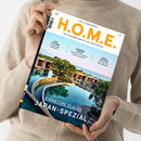 Person in beigem Strickpullover hält das Magazin „H.O.M.E. – Das Designmagazin zum Wohlfühlen“ (Ausgabe August 2025). Titelbild zeigt eine moderne Hotelanlage mit geschwungenem Infinitypool, klarer Wasseroberfläche, Bäumen und flachen Gebäuden vor hellem Himmel. Aufschrift: „SIGNATURE PLACES JAPAN-SPEZIAL“, Themen u. a. „AUF SCHIENE Luxuszeit weltweit“, „BLENDED TRAVEL Arbeiten, wo andere Urlaub machen“, „NEUE YACHTEN Wohnen auf dem Wasser“.