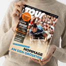Eine Person in beigem Strickpullover hält das Magazin „tour – Europas Rennrad-Magazin Nr. 1“ in den Händen, Titelbild mit einem männlichen Radrennfahrer in blauem Trikot über dem Lenker eines Rennrads. Textaufschriften: „Die besten Renner für Wettkampf & Sport“, „Teuer vs. Günstig: Rennradhelme“, „Schottland entdecken“, „Radsport in der Provinz“, „Hommage an den Größten – Zum 80. Geburtstag von Eddy Merckx“.