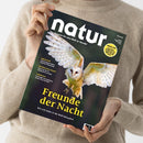 Person in hellbeigem Strickpullover hält mit beiden Händen eine Ausgabe des Magazins „natur“. Das Cover zeigt eine fliegende Schleiereule mit ausgebreiteten Flügeln vor dunkelgrünem Hintergrund. Titelaufschrift in großer gelber Schrift: „Freunde der Nacht“, Untertitel: „Wie sich Eulen in der Welt behaupten“. Weitere Texte: „Jaguarwald“, „Zertifiziertes Palmöl“, „Vogelschützer“ und gelber Kreis mit Themenhinweis oben rechts.