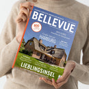 Eine Person mit beigefarbenem Strickpullover hält das Immobilienmagazin „BELLEVUE“ mit blauem Titelhintergrund und orangefarbener Randleiste, darauf ein Haus mit Reetdach, Garten und Terrasse. Sichtbare Texte: „EUROPAS GRÖSSTES IMMOBILIEN-MAGAZIN“, „Großes Special HAMBURG“, „LIEBLINGSINSEL – Sylt zwischen Sehnsucht und Realität“, „800 Angebote aus aller Welt“, sowie Themen zu Österreich, Spanien, New York und Immobilien weltweit.