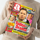 Person in beigem Pullover hält die Zeitschrift „die aktuelle“ in beiden Händen, Titelbild mit großem roten Schriftzug „Todes-Drama um seinen Vater“ und dem Namen „Julian Nagelsmann“ darunter; zudem Porträt eines Mannes in grauer Jacke mittig, kleinere Porträts zweier weiterer Personen links und rechts, gelbe und weiße Textfelder mit Schlagzeilen zu Themen wie Operation, Liebe und Prominenz, Hintergrund farbenfroh in Rot-, Gelb- und Blau-Tönen gestaltet.