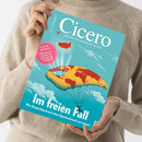 Magazin „Cicero – Magazin für politische Kultur“ in den Händen einer Person mit beigefarbenem Strickpullover; Titelblatt zeigt eine Illustration eines fallenden Erdballs mit aufgespanntem Fallschirm über blauem Himmel mit Wolken. Aufschrift: „Im freien Fall – Wie Deutschland sich dem Epochenbruch verweigert“ und runder roter Hinweis „Letztes Gefecht – Die Debatte um die AfD hält die Gesellschaft in Geiselhaft“.