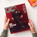 Titelbild des Magazins „GQ Germany“ in den Händen einer Person, zeigt einen männlich wirkenden Mann im schwarzen Smoking mit Fliege, sitzend vor rotem Wabenmuster-Hintergrund; weiße Textaufschriften u. a. „BEN AFFLECK – EIN BESUCH BEIM BOSS“, sowie weiter rechts Namen und Themen anderer Beiträge in weißer Schrift.