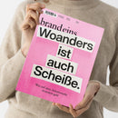 Eine Person in beigem Strickpullover hält ein Magazin mit rosa Cover in beiden Händen; auf dem Titel steht oben „brandeins“, mittig in großen schwarzen Buchstaben „Woanders ist auch Scheiße.“ und darunter kleiner „Was auf dem Arbeitsmarkt trotzdem geht“, mit sichtbarem Barcode im oberen Bereich.