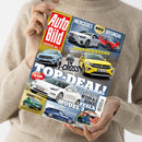 Helle, langärmlig gekleidete Person hält ein Exemplar der Zeitschrift „Auto Bild“ mit rotem Logo und gelbem Rand. Auf dem Titel sind mehrere Autos abgebildet, darunter VW T‑Cross, Tesla Model 3, Mercedes C‑Klasse, Hyundai Ioniq 3, Renault Twingo und BMW M340i Touring. Sichtbare Schlagworte: „Kaufberatung“, „Top‑Deal!“, „Gebraucht besser als gedacht“ und „Europas Nr. 1“.
