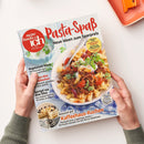 Titelbild der Zeitschrift „meine Familie & ich“ mit dem Hauptschriftzug „Pasta-Spaß – Neue Ideen zum Sparpreis“. Abgebildet ist ein Teller Nudeln mit Hackfleischsoße, Kirschtomaten, Kräutern und einem Löffel und einer Gabel auf blau-weiß karierter Tischdecke. Weitere Texte: „Kaffeehaus-Kuchen“, „Vegetarisch feiern“, „Hähnchen-Hits“, „Spargel aus dem Ofen“ und „Geling-Garantie“. Gehalten von zwei Händen vor hellem Hintergrund.