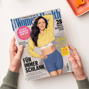 Titelbild der Zeitschrift „Women’s Health“ mit einer lächelnden weiblichen Person in gelbem Cropped-Hoodie und blauer Sportshorts vor beigem Hintergrund. Textelemente: „FÜR IMMER SCHLANK“, „28 Tricks, besser zu essen“, „Effektiv trainieren ohne Gym“, „Dein Weg zum Super-Bauch“, Name „Jasmin Wagner“ und Markenname „Fabletics“ auf dem weißen Sporttop. Zwei Hände halten das Magazin über eine helle Oberfläche mit orangefarbenem Tablett.
