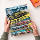 Titelbild der Zeitschrift „Auto Bild sportscars“ in den Händen einer Person mit grünem Strickpullover, gezeigt vor hellem Hintergrund. Das Cover zeigt oben ein gelb-schwarz getarntes Sportwagenmodell mit der Überschrift „Lamborghini Temerario – Erste Fahrt im Huracán-Nachfolger“. Darunter ein roter Mercedes-AMG GT 63, mehrere kleinere Autos und Textfelder zu Tests, Vergleichen und Kaufberatung.