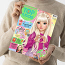 Person mit beiger Strickjacke hält ein Magazin mit dem Titel „TOP Model“ in hellgrün-blauer Schrift vor sich; auf dem Cover eine gezeichnete Figur mit langen, blonden Haaren, pinkem Pullover und Händen zu einem Herz geformt. Um die Figur bunte Textfelder mit Aufschriften wie „Knusper-Frühstück“, „Ein unvergesslicher Geburtstag“, „Happy Ostern“ und „Stylecheck“, dazu kleine Bilder, Sterne und QR-Code.