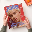 Titelbild der Zeitschrift „jolie“ mit großem violettem Logo oben auf rotem Hintergrund; im Mittelpunkt das Porträt einer weiblich gelesenen Person mit kurzem blondem Haar. Textausschnitte: „Die neuen Trend-Labels“, „Streetstyles aus der ganzen Welt“, „Stehen mir kurze Haare?“, „Die beste Investition deines Lebens“. Zwei Hände halten das Magazin über einer weißen Tischoberfläche mit orangefarbenem Behälter am Rand.