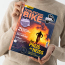 Person in beigefarbenem Strickpullover hält ein Magazin mit dem Titel „mountain BIKE – ALLES IST FAHRBAR“. Cover zeigt eine Silhouette eines Mountainbikers vor orange-gelbem Sonnenuntergang. Textelemente u. a.: „Der PREIS ist HEISS“, „21 Enduro- & Trail-Reifen im Test“, „Orbea – Das neue E-MTB-Flaggschiff“, „ZELL AM SEE“, „Shimano XT Di2 im Stresstest“ und „Neu – 16 Seiten Community“.
