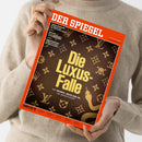Person in beigefarbenem Strickpullover hält ein Exemplar des Magazins „DER SPIEGEL“ mit rotem Rand und braunem Cover im Hochformat; mittig steht in großen gelben Buchstaben der Titel „Die Luxus-Falle – Wie Hermès, Rolex und Co. uns manipulieren“, Hintergrund mit goldfarbenen Symbolen wie Kronen, Blumen, Totenkopf und Schlange auf lederähnlicher Struktur.