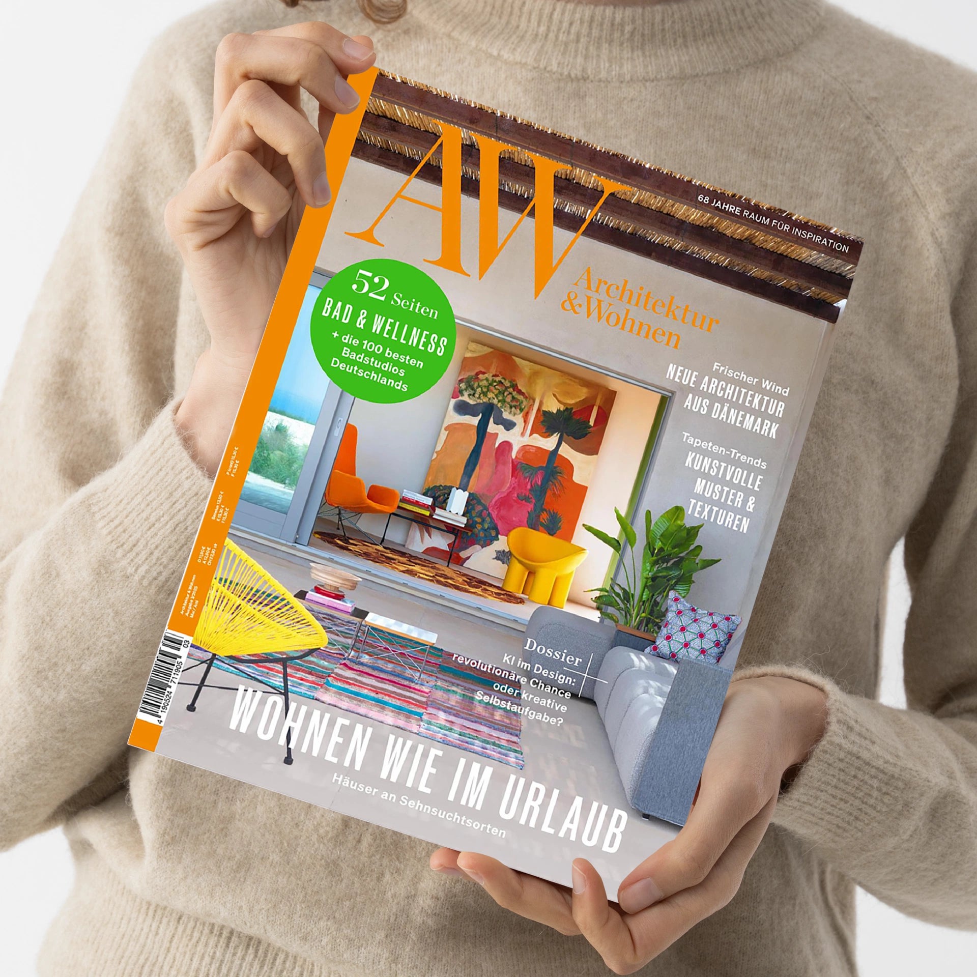 Person in beigem Strickpullover hält die Zeitschrift „AW Architektur & Wohnen“ mit orange-weißer Titelleiste und großem Innenraumbild mit bunter moderner Einrichtung. Sichtbare Texte: „52 Seiten Bad & Wellness – die 100 besten Badstudios Deutschlands“, „Wohnen wie im Urlaub – Häuser an Sehnsuchtsorten“, „Neue Architektur aus Dänemark“, „Kunstvolle Muster & Texturen“.