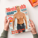Titelbild der Zeitschrift „Men’s Health“ mit großem roten Schriftzug oben; im Mittelpunkt ein lächelnder, muskulöser Mann mit nacktem Oberkörper, dunkler Badehose und Surfbrett am Strand vor blauem Himmel. Textaussagen u. a.: „DEIN BESTER SOMMER“, „MIT 50 FITTER ALS MIT 35!“, „Einfach genial leben mit JAMIE OLIVER“, „Verstehst du die Frauen?“ und „Der total ehrliche Alkohol-Test“. Hände halten das Heft über heller Tischfläche.