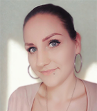 Nahaufnahme einer weiblichen Person mit zurückgebundenem dunklem Haar, hellen Augen und dezent geschminktem Gesicht mit betonten Augenbrauen und Eyeliner; sie trägt große silberfarbene Creolen, eine feine Kette, ein Oberteil in zartem Rosaton mit V-Ausschnitt sowie ein kleines Piercing unterhalb der Unterlippe, vor einem einfarbig hellen Hintergrund mit weichem Lichteinfall von der Seite.
