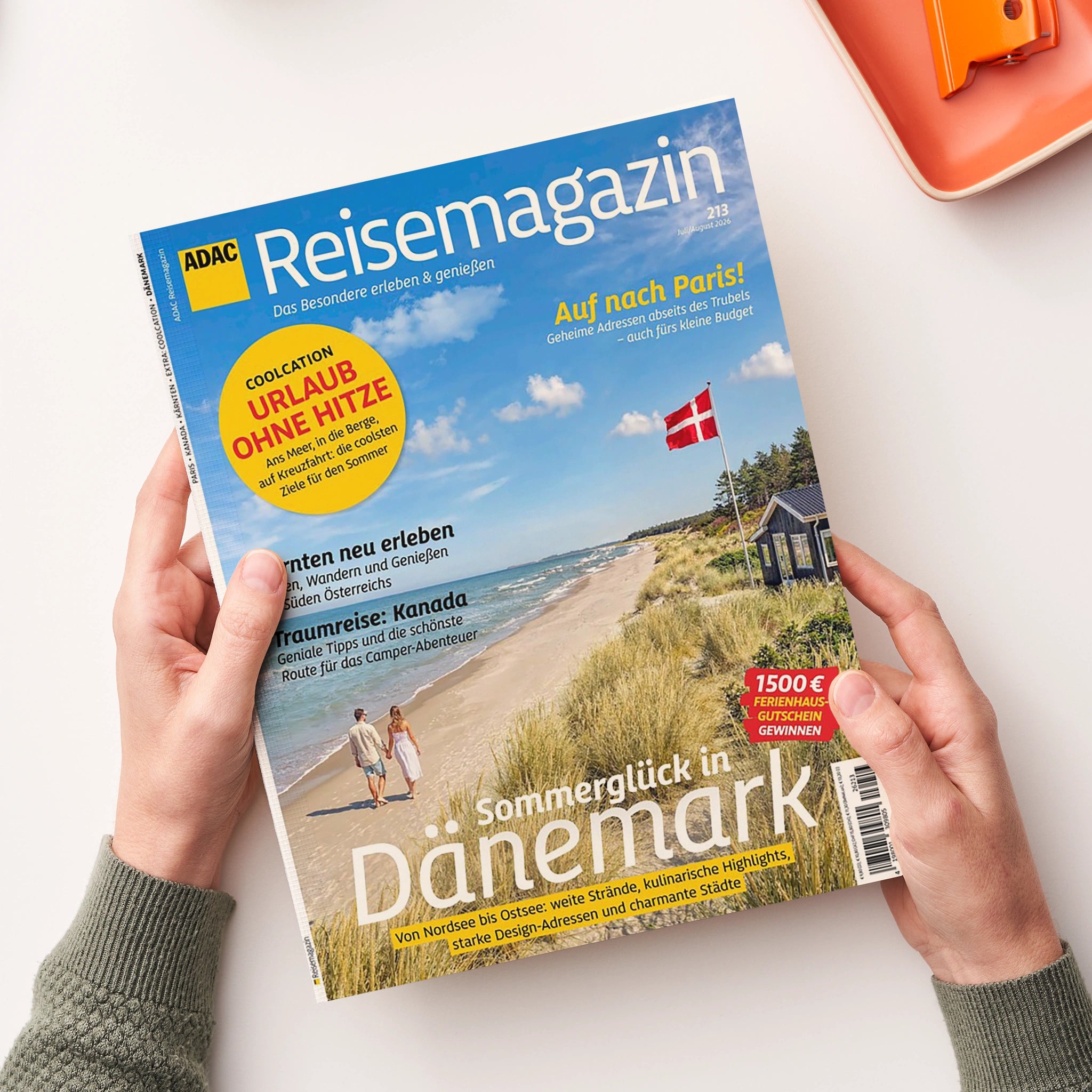 Magazintitel „ADAC Reisemagazin“ mit blauem Hintergrund und Foto einer Küstenlandschaft mit Dünen, Sandstrand, Meer und Haus mit dänischer Flagge; im Vordergrund zwei gehende Personen. Gelbe, blaue und weiße Überschriften unter anderem: „Sommerglück in Dänemark“, „URLAUB OHNE HITZE“, „Traumreise: Kanada“ und „Auf nach Paris!“. Gehalten von zwei Händen vor hellem Hintergrund, rechte obere Ecke der Seite leicht angehoben.