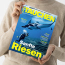 Beige gekleidete Person hält die Zeitschrift „TAUCHEN – Europas größte Tauchzeitschrift“ mit blauem Cover, auf dem ein Taucher in schwarzer Ausrüstung und gelben Flossen neben einem Mantarochen unter Wasser zu sehen ist; sichtbare Überschriften: „Sanfte Riesen – Alles Wissenswerte über Mantas“, „Reisewelten: Dänemark, Malediven, Ost-Timor“, „Medizin: Tauchen mit Diabetes“, „Tests & Tipps: Unterzieher und Karabiner“.