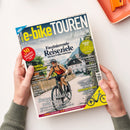 Titelbild des Magazins „e-bike TOUREN“ mit dem Untertitel „Faszinierende Reiseziele mit dem E-Bike entdecken“, gehalten von zwei Händen über hellem Tisch, daneben orangefarbene Schale. Auf dem Cover eine Person mit rotem Trikot, schwarzer Hose und Helm auf einem E-Bike vor alpiner Dorflandschaft. Farbtöne in Gelb, Blau und Weiß, mit Texten u. a. „30 Reisetipps“, „Berge, Wälder und Seen“ und „E-Bike Highlights 2025“.