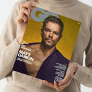 Person in beigefarbenem Wollpullover hält eine Ausgabe des Magazins „GQ Germany“. Auf dem Cover ist ein kurzhaariger, bärtiger Mann mit geöffnetem dunkelviolettem Hemd vor gelbem Hintergrund abgebildet. Sichtbare Textelemente: „DAS MAKING OF DIE ODYSSEE MIT MATT DAMON“, „TOM HOLLAND & ROBERT PATTINSON“, „PLUS“ sowie weitere kleinere Überschriften zu Themen wie Uhren, Fußball-WM und Pride-Special.