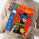 Person in beigem Pullover hält das Kindermagazin „ZEIT Leo“ mit rotem Titelblatt, darauf eine Ziege mit blauer Superheldenmaske und -umhang vor gelbem Sternenmuster. Hauptüberschrift „Die Superkraft der Tiere“, weitere sichtbare Texte: „WM-Stars“, „Spurensuche“, „Tempo 300“ sowie Logoillustration einer Comicfigur und der Schriftzug „DIE ZEIT“ auf blauem Seitenstreifen.