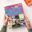 Titelblatt des Magazins „Bastei Glücks Rätsel“ mit pinkfarbenem Hintergrund und großer blauer Schrift. Im oberen Bereich steht „Der große Freizeitspaß“, unten sind Fotos mit möglichen Gewinnen wie Tierpflege-Sets, Frühstücks-Sets, Olivenöl-Sets, Gläser-Sets, Freizeitpark-Tickets und Urlaub abgebildet. Links ist eine lächelnde Frau mit blondem Haar gezeigt, rechts oben ein Rätselgitter auf gelbem Feld. Zwei Hände halten das Heft.