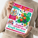 Helle, beige gekleidete Person hält das Magazin „Good Health“ in den Händen; auf dem farbenfrohen Titelbild sind Spinatblätter, halbierte Kirschtomaten, Blaubeeren, Walnüsse und Mandeln abgebildet. Texte u. a.: „Endlich wieder neue Kraft tanken“, „Wie Bewegung unser Gehirn fit hält“, „So erkennen wir unsere Schutz-Strategien“ und „Wie Tomaten und Blaubeeren unseren Zellen frische Energie schenken“.