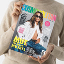 Hellhäutige Person in beigem Strickpullover hält eine Ausgabe der Zeitschrift „COSMOPOLITAN“ (Juni 2025) vor grauem Hintergrund, Cover in kräftigen Farben mit einer weiblichen Person in aufgeknöpfter weißer Bluse, Jeans und Sonnenbrille. Sichtbare Texte: „MUT IST MEIN MUSKEL“, „LET LOVE RULE“, „SIE SIND REICHER, ALS SIE GLAUBEN“, „BULLSHIT? BINGO“, „KOMM WAHNER“.