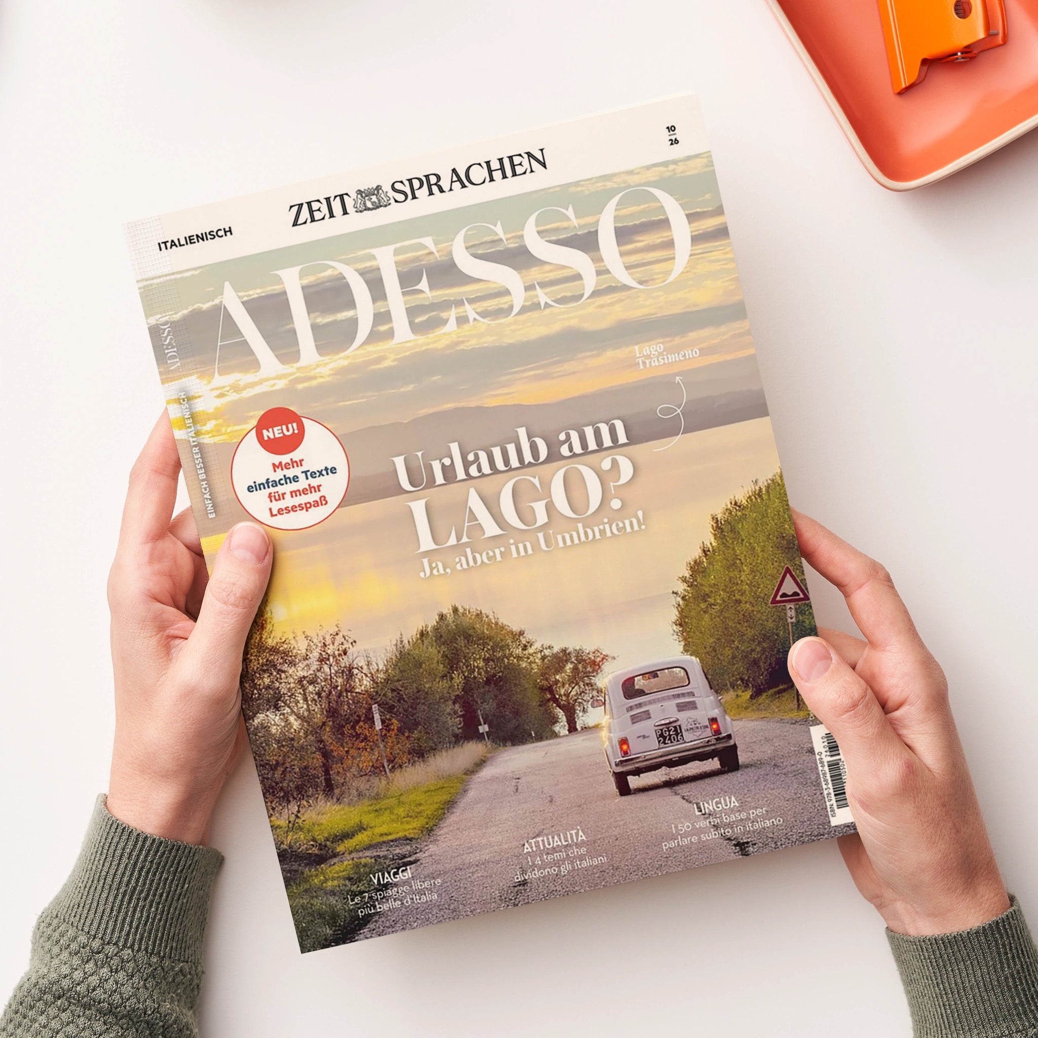 Aufgeschlagenes italienisches Sprachmagazin „ADESSO“ von „ZEIT SPRACHEN“ auf weißer Oberfläche, von zwei nicht näher bestimmbaren Händen gehalten. Titelzeilen: „ITALIENISCH“, „Urlaub am LAGO? Ja, aber in Umbrien!“, „Logo Trasimeno“ und „NEU! Mehr einfache Texte für mehr Lesespaß“; darunter Landschaftsstraße mit weißem Auto, Bäumen und Verkehrsschild. Im Hintergrund liegt ein orangefarbenes Bürogerät.