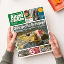 AngelWoche, eine deutschsprachige Sportfischer-Zeitung, wird von zwei Händen über einer weißen Fläche gehalten. Das Cover zeigt grüne und blaue Gestaltung, mehrere Fische im Wasser sowie die Texte „Deutsche Sportfischer Zeitung“, „Angel Woche“, „Das große Fressen!“, „Barsch frisst Larve“, „Hecht frisst Barsch“, „Angler isst Hecht“, „Frage der Woche“ und „Wie gelingt das Lockfutter perfekt?“.