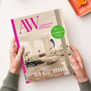 Von zwei Händen gehaltenes, schräg liegendes Magazin „AW Architektur & Wohnen“ mit cremefarbenem Cover, pinkem Schriftzug und Foto eines hellen Wohnzimmers. Sichtbare Texte: „69 Jahre Raum für Inspiration“, „36 Seiten Spezial Ferienimmobilien Einrichten & vermieten“, „Der neue Norden“ und „Design-Highlights und Interior-Ideen aus Skandinavien“; oben rechts liegt teilweise ein orangefarbenes Objekt.