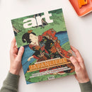 Aufgeschlagenes Kunstmagazin „art“ in schräger Draufsicht, von zwei Händen über weißer Fläche gehalten; Cover mit farbiger japanischer Holzschnittdarstellung einer Frau im dekorierten Kimono vor grüner Landschaft und blauem Wasser. Sichtbare Texte: „DAS KUNSTMAGAZIN // AUGUST 2026“, „JAPANLIEBE“, „MIT POSTER-EDITION ZUM HERAUSNEHMEN!“, „MANIFESTA 16 RUHR“ und „PAUL SIGNAC IN POTSDAM“.