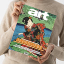 Kunstmagazin „G...“ mit grün-blauem Cover und japanischer Holzschnittdarstellung einer Frau im floral gemusterten Kimono, gehalten von einer Person im hellbeigen Strickpullover. Sichtbare Texte: „DAS KUNSTMAGAZIN/AUGUST 2026“, „JAPAN LIEBE“, „MIT POSTER-EDITION ZUM HERAUSNEHMEN!“, „MANIFESTA 16 RUHR“ und „PAUL SIGNAC IN POTSDAM“.
