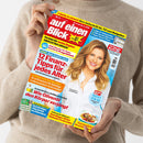Hände einer nicht näher bestimmbaren Person halten schräg eine farbige Zeitschrift vor einem beigen Strickpullover. Das Cover trägt „auf einen Blick“, zeigt eine blonde Frau im weißen Hemd und enthält Texte wie „2 Hefte in einem! Ihre Zeitschrift für TV und Ratgeber“, „12 Finanz-Tipps für jedes Alter“, „SWR EXTRA GROSS!“ und „Wie Olivenöl den Körper verjüngt“.