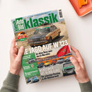 Schräg auf einer weißen Fläche liegende Ausgabe der Zeitschrift „Klassik Auto Bild“, gehalten von zwei Händen mit grünen Strickärmeln; auf dem Cover ein beigefarbener Mercedes-Benz W123 mit Dachgepäck auf einer Wüstenstraße, ergänzt durch eine marokkanische Flagge und mehrere Oldtimerbilder. Sichtbare Texte: „W123 Challenge“, „Die Jagd auf W123“ und „Im alten Benz quer durch Marokko“; oben rechts liegt ein orangefarbenes Tablett.